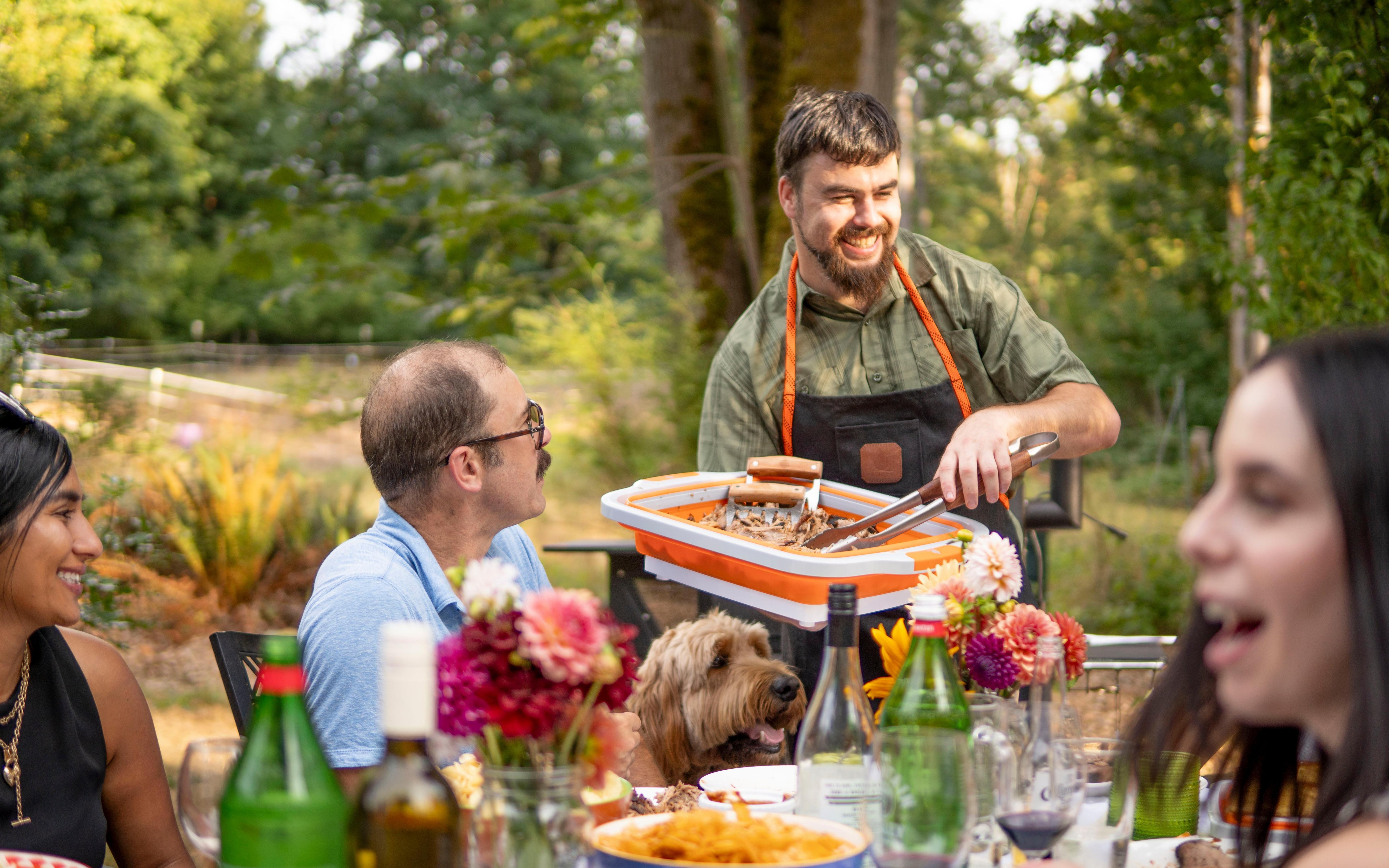 Man serving food with tongs at a table. There is a group of friends and a dog outdoors with drinks and flower centerpiece.